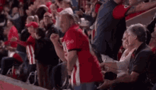 a man in a red shirt is jumping in the air while sitting in the stands at a basketball game .