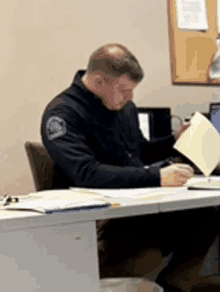 a man in a black jacket is sitting at a desk holding a piece of paper .