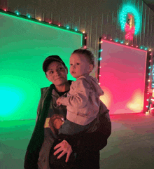 a woman is holding a baby in front of a wall with christmas lights on it