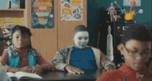 a boy with a white mask on his face is sitting at a desk in a classroom with other children .