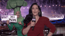 a woman holding a sign that says hi mom stands next to a woman in a broccoli costume