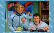 two men are sitting at a desk in front of a superman poster and a book .