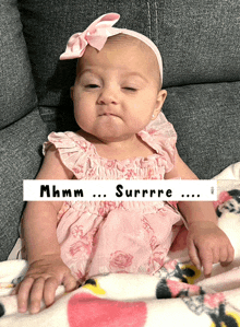 a baby in a pink dress is sitting on a couch with a sign that says mhmm surrre