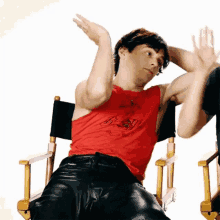 a man in a red tank top and leather pants is sitting in a director 's chair with his hands in the air .