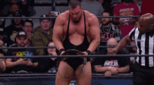 a wrestler is standing in a ring with a referee and a crowd behind him with a shirt that says american