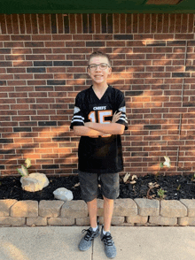 a young boy wearing a black chiefs jersey stands with his arms crossed