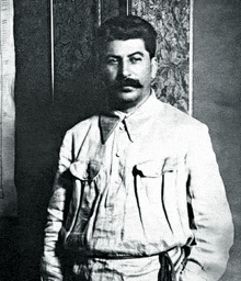 a black and white photo of a man with a mustache wearing a white shirt and standing in front of a wall .