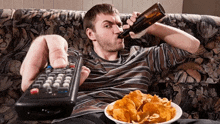 a man sits on a couch drinking a beer and eating chips