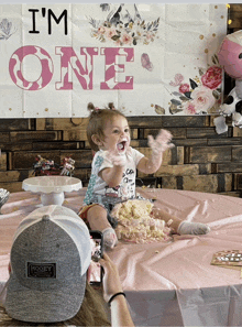 a baby is sitting at a table with a cake and a sign that says i 'm one