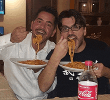 two men are eating spaghetti next to a bottle of coca-cola