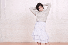 a woman wearing a grey sweater and a white skirt stands in front of a white wall