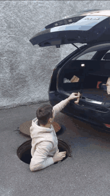 a man is sticking his head out of a manhole cover in front of a car with the trunk open