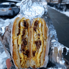 a person is holding a sandwich in aluminum foil with a car in the background