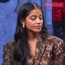 a woman with long black hair is wearing a floral jacket and earrings and is sitting in front of a wall .