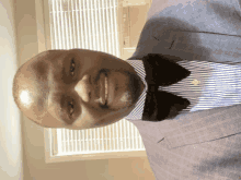 a man in a suit and bow tie smiles in front of a window with blinds