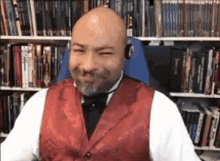 a bald man with a beard wearing a red vest and bow tie is sitting in a chair in front of a bookshelf .