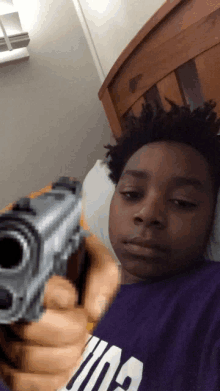 a boy in a purple shirt is laying on a bed with a gun in the background