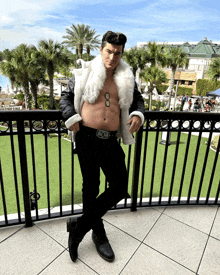 a shirtless man leans against a railing wearing a black jacket and a white fur vest