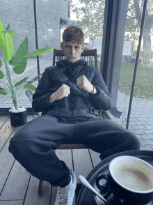 a man sits in a chair with his legs crossed and a cup of coffee in front of him