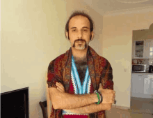 a man with a mustache and a scarf around his neck is standing with his arms crossed in a living room .