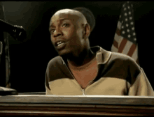 a bald man is sitting at a desk in front of a microphone with an american flag in the background .