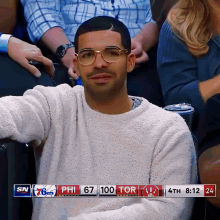 a man wearing glasses sits in the stands watching a basketball game with the scoreboard showing philadelphia 67 100 tor