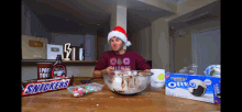 a man wearing a santa hat is sitting at a table with a bowl of food and a box of oreo cookies