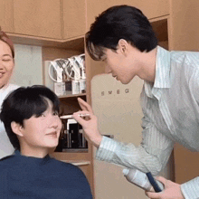 a man is applying makeup to another man 's face in a kitchen in front of a smeg refrigerator .