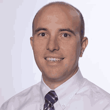 a bald man in a white shirt and purple tie smiles for the camera