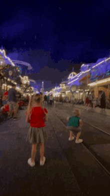 a little girl in a red shirt and a little boy in a blue shirt are walking down a street at night