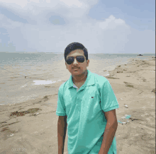 a young man wearing sunglasses and a teal shirt stands on a beach