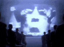 a group of people are standing in front of a screen with a picture of a man 's face projected on it .