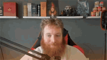 a man with a beard is sitting in front of a microphone in front of a shelf with books on it