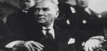 a black and white photo of a man in a suit and tie sitting in a crowd of people .