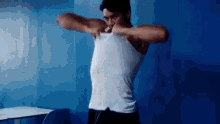 a man in a white tank top is standing in front of a blue wall and taking off his shirt .