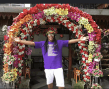 a man in a purple shirt stands in front of a floral arch