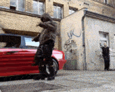 a man in a hoodie stands in front of a red car with graffiti on the wall that says uk