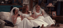 three women in wedding dresses are sitting on a couch drinking and eating popcorn