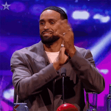 a man in a suit is clapping his hands while sitting at a table in front of a microphone .