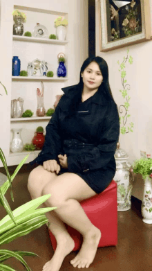 a woman in a black coat sits on a red ottoman in front of shelves full of vases