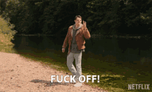 a man in a brown jacket is giving the middle finger in front of a body of water and the word fuck off is written below him