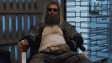 a man with a beard and sunglasses is sitting in a chair holding a drink .