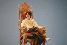 a man in a white shirt sits on a gold throne with a red cushion