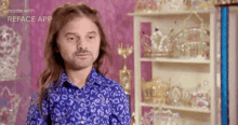 a man with long hair and a beard wearing a blue shirt is standing in front of a shelf full of trophies .