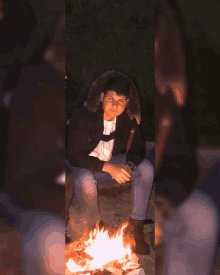 a boy in a black jacket sits in front of a fire at night