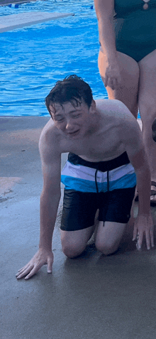 a boy in swim trunks is kneeling on the ground near a swimming pool