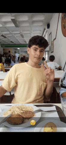 a young man sitting at a table with a plate of food giving the peace sign