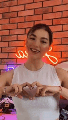 a woman in a white tank top is making a heart with her hands in front of a brick wall .