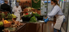 a woman wearing a mask and blue gloves is standing in front of a table with vegetables on it .