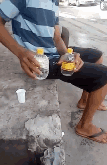 a man in a striped shirt sits on a bench holding two bottles of water and a cup of water